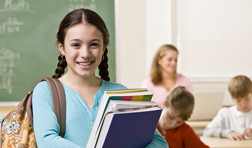 Middle school girl with dark hair stands in a classroom, holding books and smiling